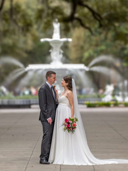 Elopement Wedding A bride and groom stand gazing at each other. The bride wears a flowing white dress and veil, holding a bouquet of red and pink flowers. The groom is in a dark suit and tie. Behind them, a large white tiered fountain is visible, with trees framing the background. Elopements Inc