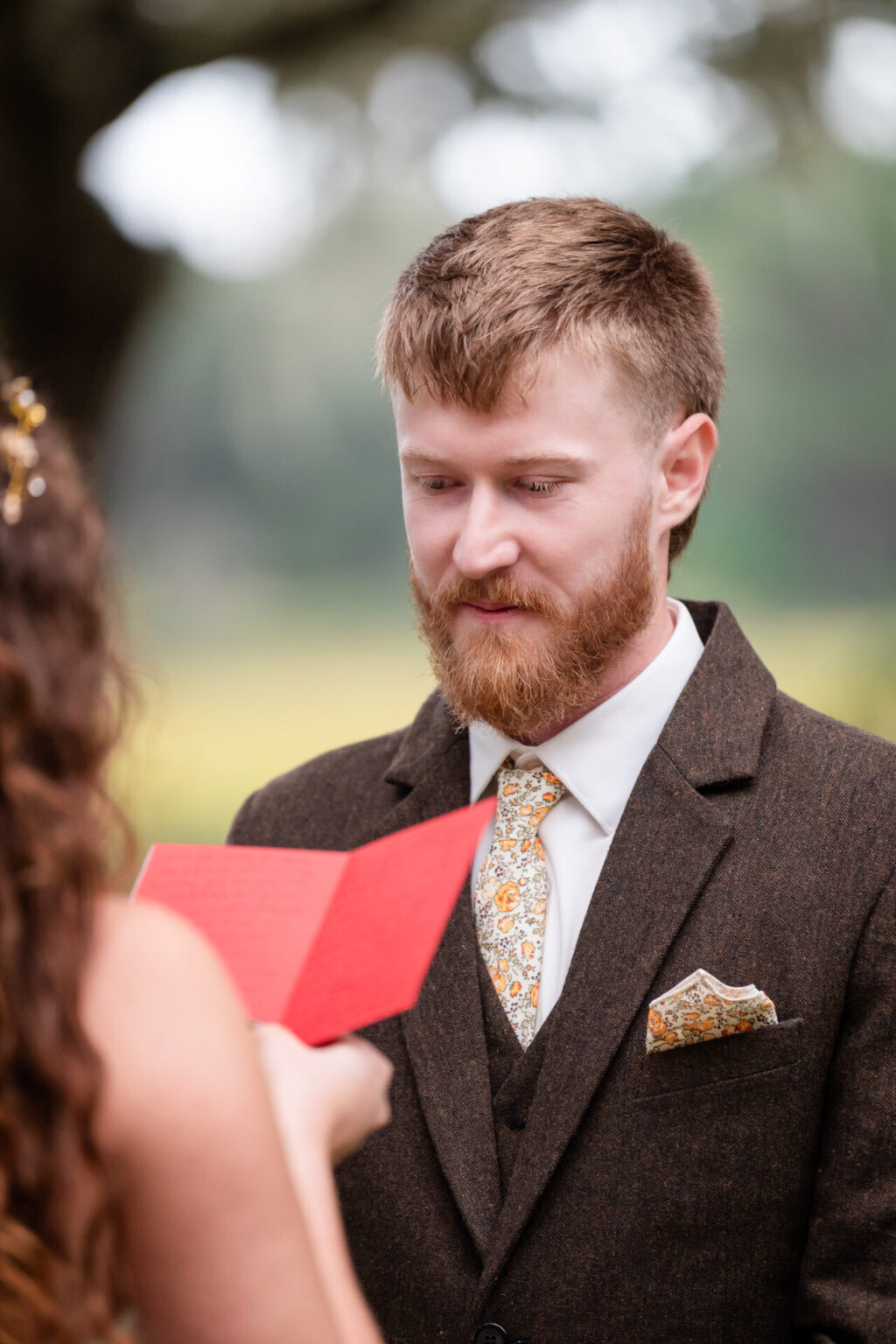 Elopement Wedding A man with a beard stands outdoors, wearing a brown suit with a floral tie and pocket square. He is looking at a red card held by a woman with long, wavy hair. The background is blurred, featuring greenery and suggesting a serene, natural setting. Elopements Inc