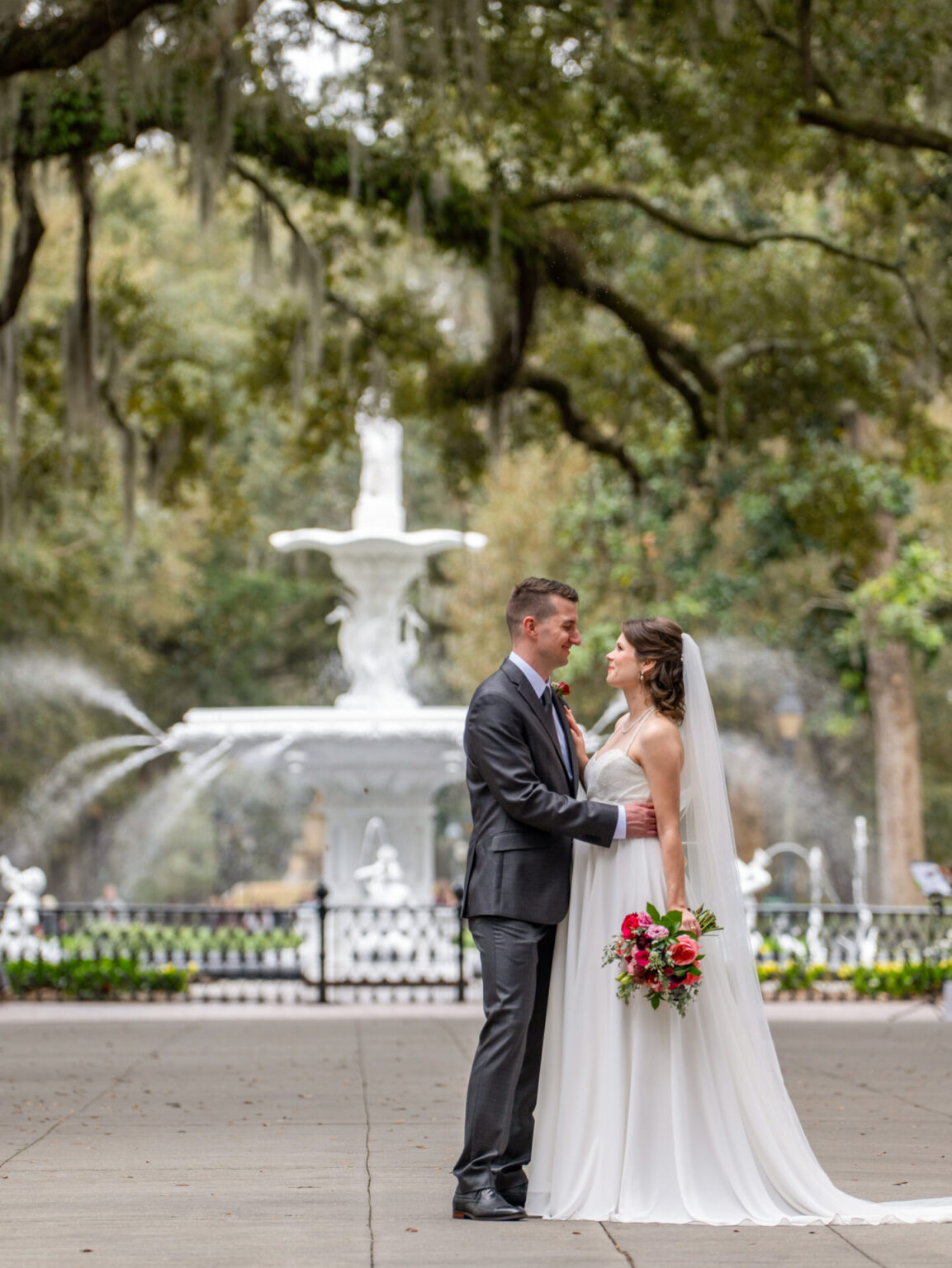 Elopement Wedding A bride and groom stand facing each other under lush trees with Spanish moss. The bride, in a white gown and veil, holds a bouquet of red and pink flowers. The groom wears a dark suit with a purple tie. Behind them, a grand white fountain is visible, framed by greenery. Elopements Inc