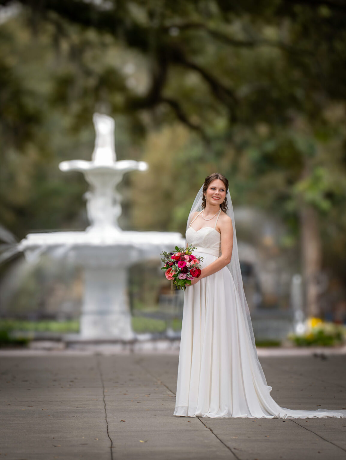 Elopement Wedding A bride in a flowing white gown stands outdoors, holding a vibrant bouquet of pink and red flowers. She smiles while looking into the distance. A large, ornate fountain is blurred in the background, surrounded by lush green trees, creating a serene and elegant setting. Elopements Inc