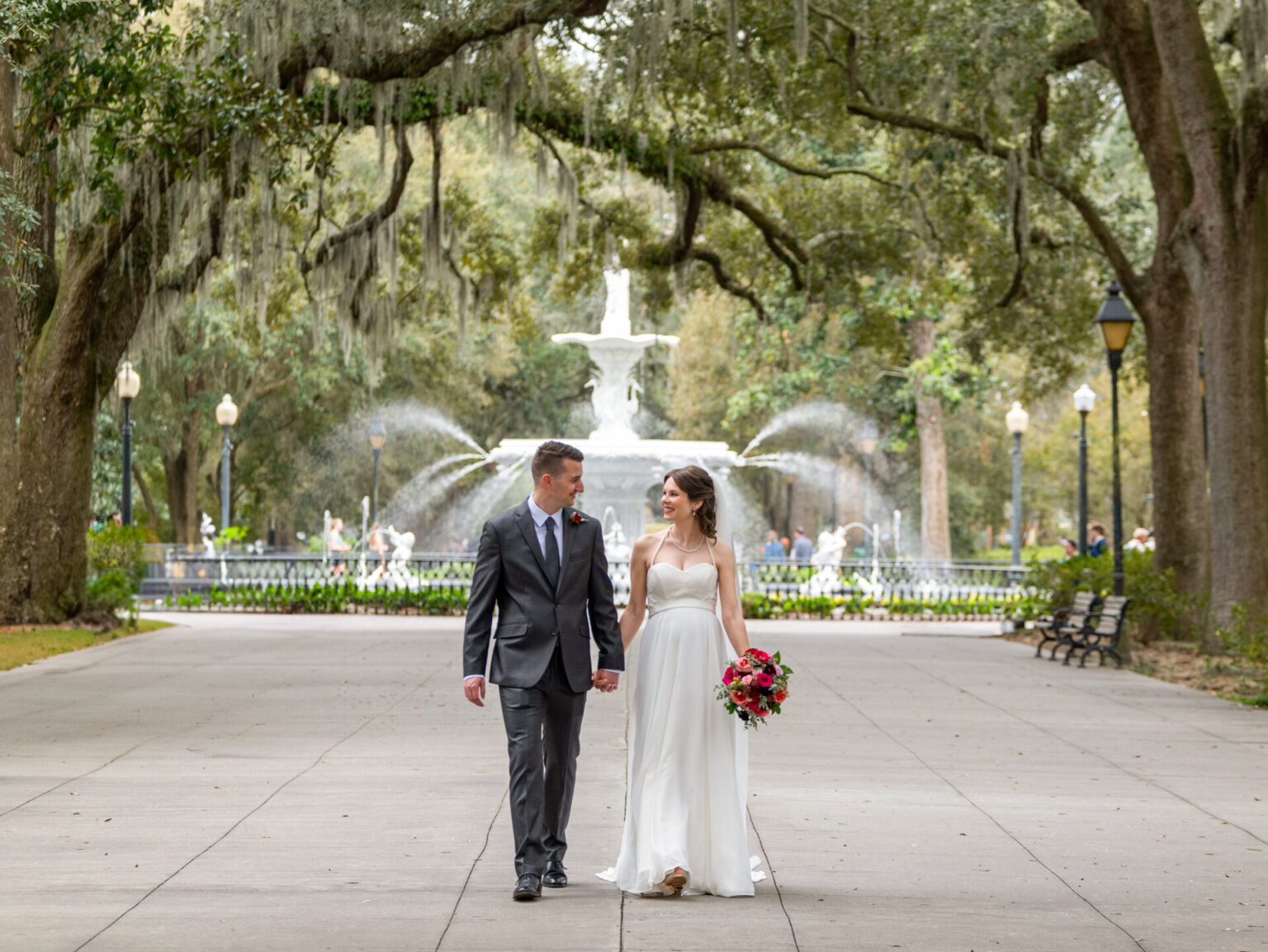 Elopement Wedding A couple walks hand in hand down a paved path in a park. The groom wears a dark suit and tie, and the bride wears a white strapless gown, holding a bouquet of red and pink flowers. Behind them, a grand fountain and lush, arching trees create a romantic backdrop. Elopements Inc