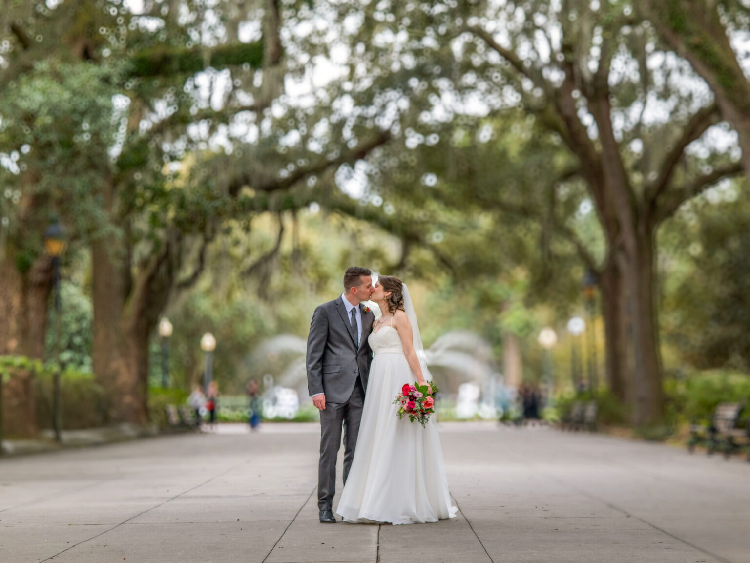 Elopement Wedding A bride and groom share a kiss on a tree-lined path. The bride wears a white gown and holds a bouquet with pink and red flowers. The groom is in a gray suit. The background features large trees and distant, blurred figures. The scene is set in an outdoor park. Elopements Inc