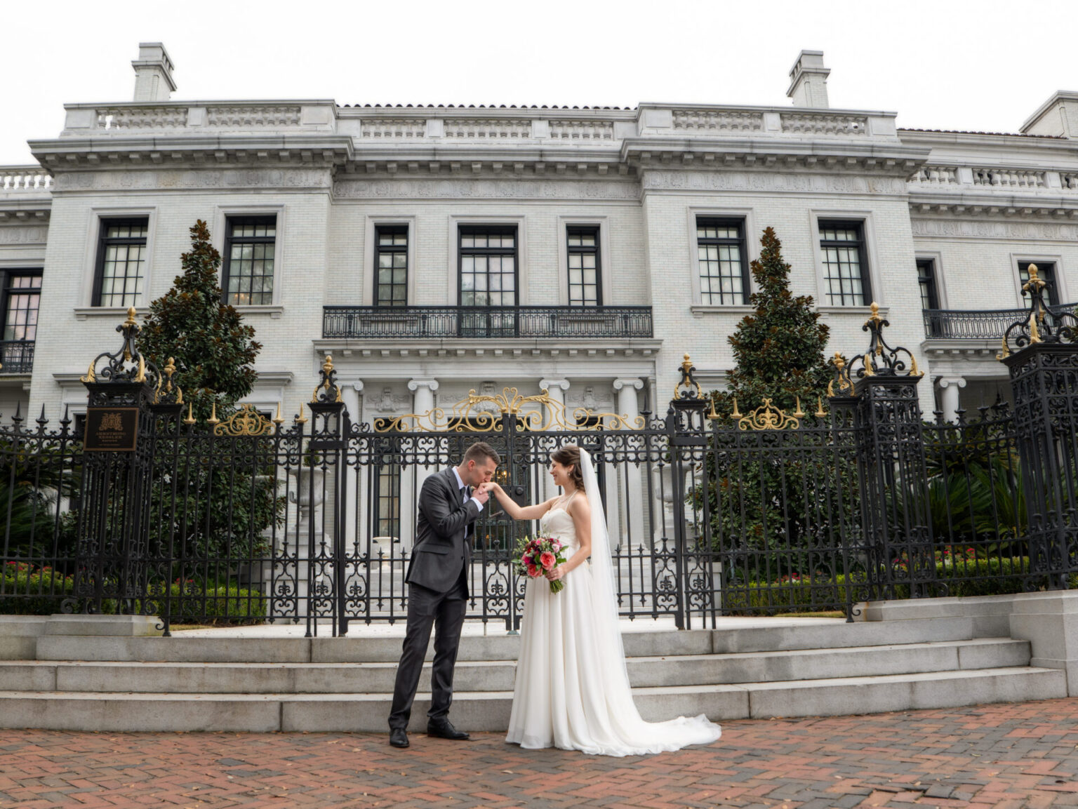 Elopement Wedding A groom in a gray suit kisses the hand of a bride in a white gown holding a bouquet. They stand in front of an ornate black gate and a grand, historic white building with tall windows and decorative elements. The brick pathway adds to the elegant setting. Elopements Inc