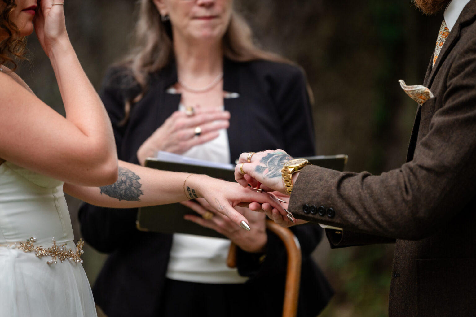 Elopement Wedding A bride and groom exchange rings during a wedding ceremony. The bride wears a white dress with floral tattoos visible on her arm, and the groom, in a brown suit, has tattoos on his hand. An officiant stands behind them, holding a book, while the bride appears emotional. Elopements Inc