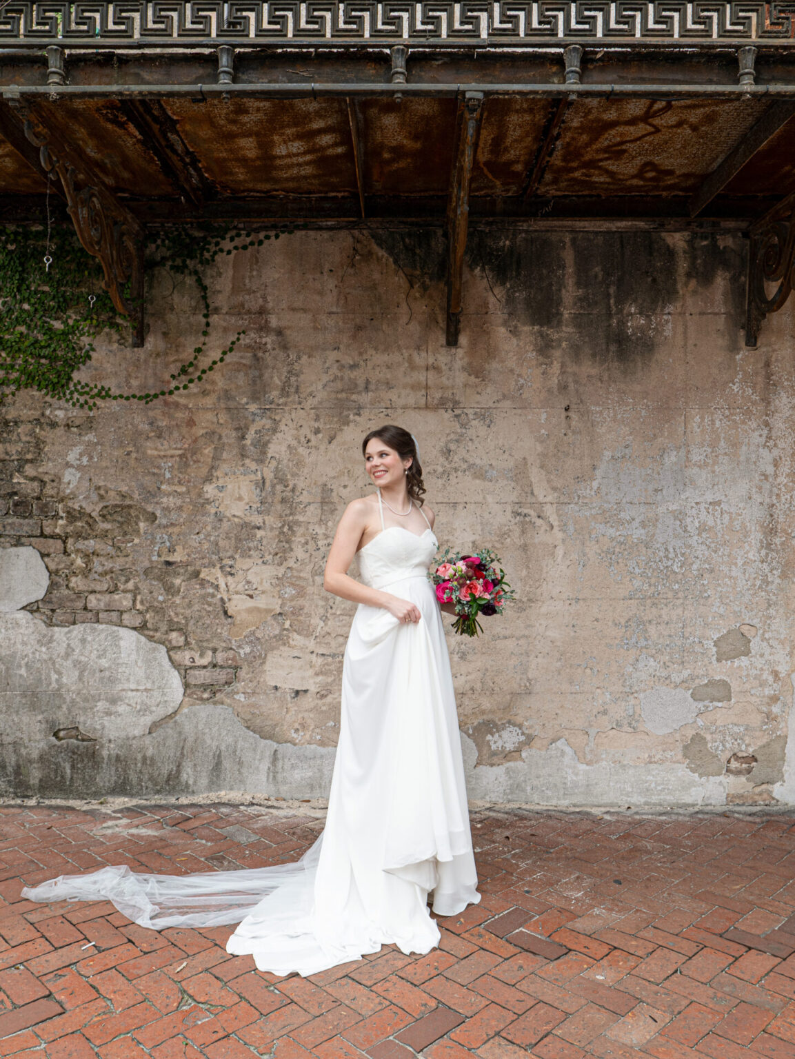 Elopement Wedding A bride in a flowing white gown stands on a brick pathway, holding a vibrant bouquet of pink and red flowers. She poses against an aged wall with ivy and intricate metalwork above. Her gown has a long train, and her hair is styled elegantly. The scene is calm and romantic. Elopements Inc