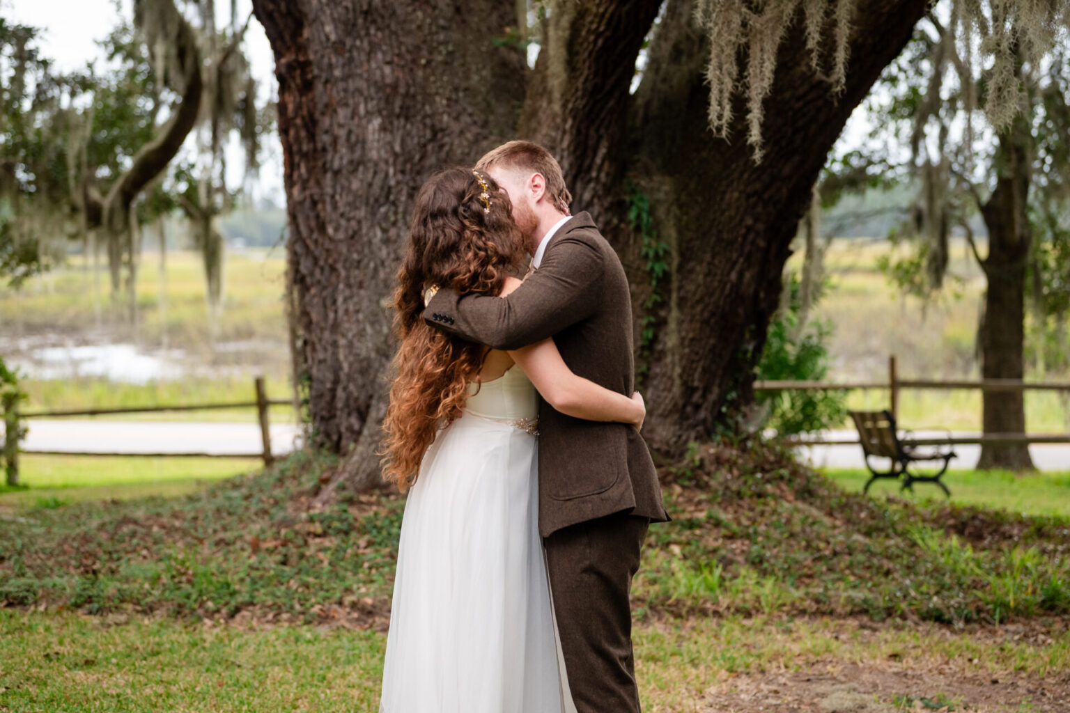 Elopement Wedding A couple is kissing in front of a large tree draped with Spanish moss. The woman, in a white dress, has long, curly brown hair. The man, in a brown suit, embraces her. A wooden fence and bench are visible in the background, with a scenic view of trees and a body of water. Elopements Inc