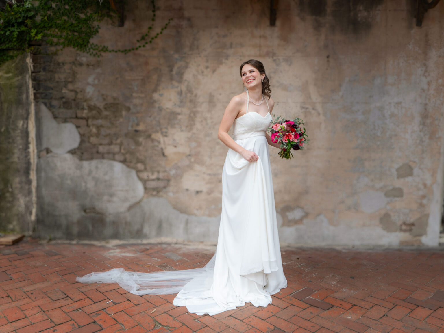 Elopement Wedding A bride in a white strapless gown stands smiling against an aged, textured wall with ivy. She holds a vibrant bouquet of pink and red flowers. The brick floor adds rustic charm, while her long veil trails behind, enhancing the romantic and elegant atmosphere. Elopements Inc