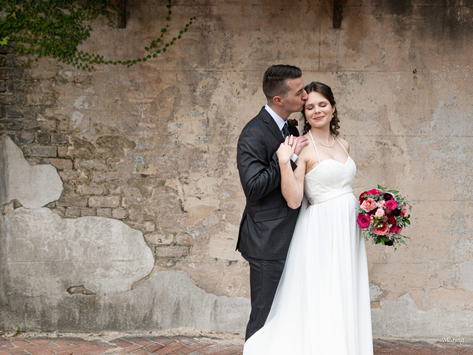 Elopement Wedding A bride and groom stand in front of an aged, textured brick wall. The groom, in a dark suit, kisses the bride's temple. The bride, in a white strapless gown, holds a bouquet of red and pink flowers. She looks content, eyes closed, while holding his hand delicately. Elopements Inc