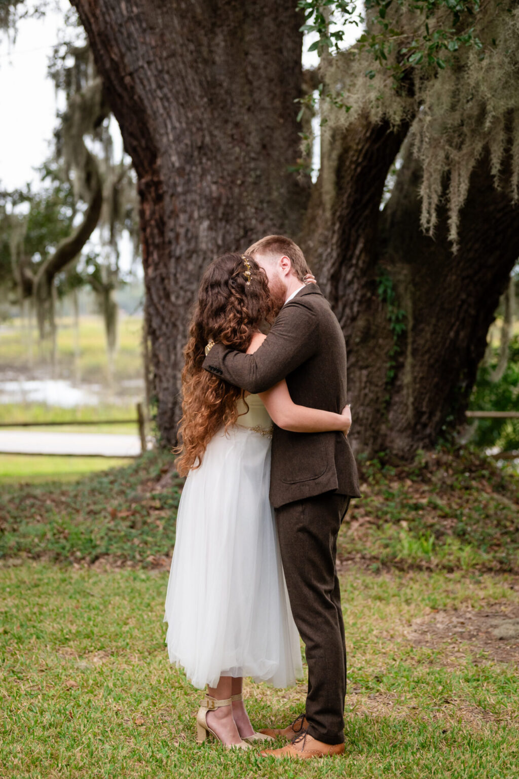 Elopement Wedding A couple embraces under a large, moss-covered tree. The woman wears a white dress and has long, curly hair, while the man is in a brown suit. They stand on green grass with a scenic, blurred background of trees and a fence, creating a serene outdoor setting. Elopements Inc