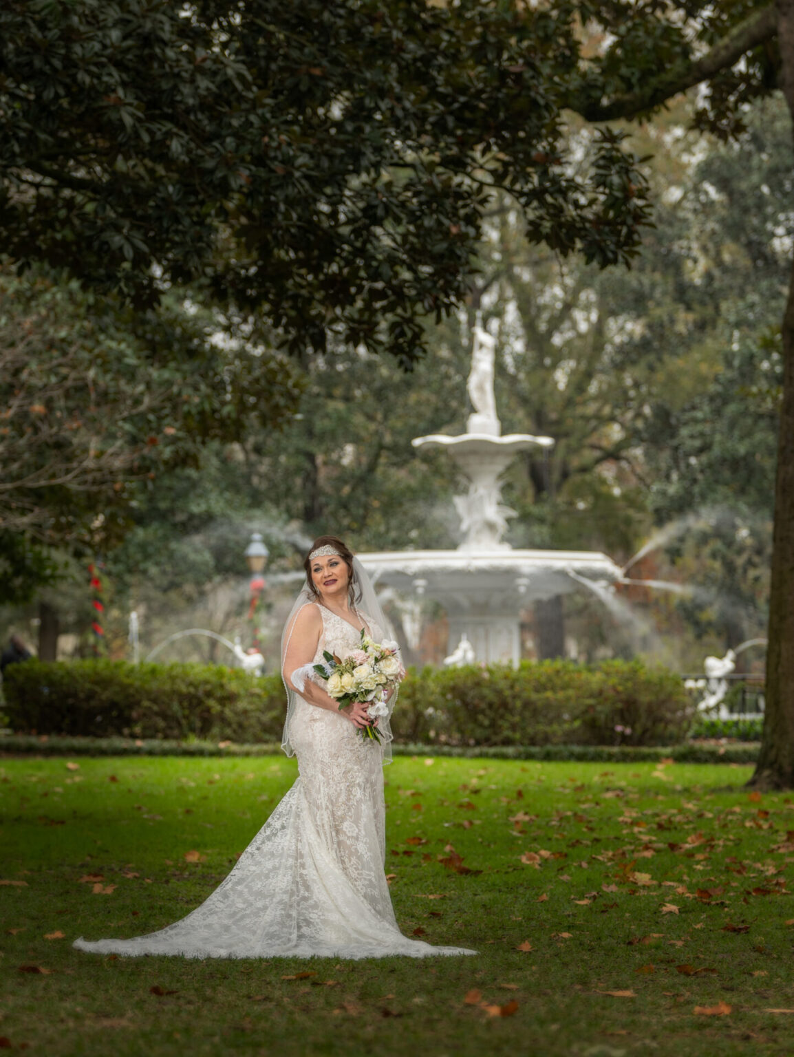 Elopement Wedding A bride in a white lace wedding dress and veil stands smiling on a grassy area in front of a fountain. She holds a bouquet of flowers and is surrounded by trees with scattered leaves on the ground. The fountain behind her has two tiers with a statue on top. Elopements Inc