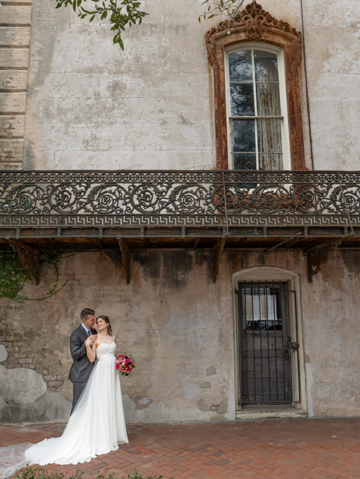 Elopement Wedding A groom in a gray suit tenderly embraces a bride in a white gown holding a bouquet of pink and red flowers. They stand in front of an aged, rustic building with a tall, ornate window and wrought iron balcony. A small, arched door is visible on the right, set in a textured, weathered wall. Elopements Inc