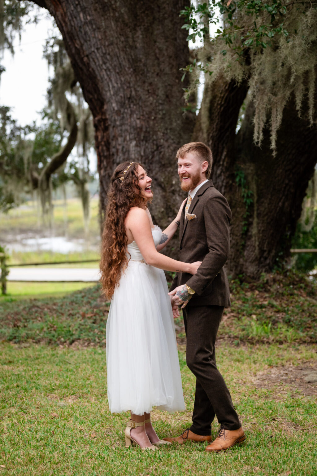 Elopement Wedding A happy couple stands outdoors in front of a large tree. The woman, with long curly hair in a white dress, smiles widely while holding hands with the man, who has a beard and wears a brown suit and tan shoes. Moss-draped branches and green grass create a serene, natural setting. Elopements Inc