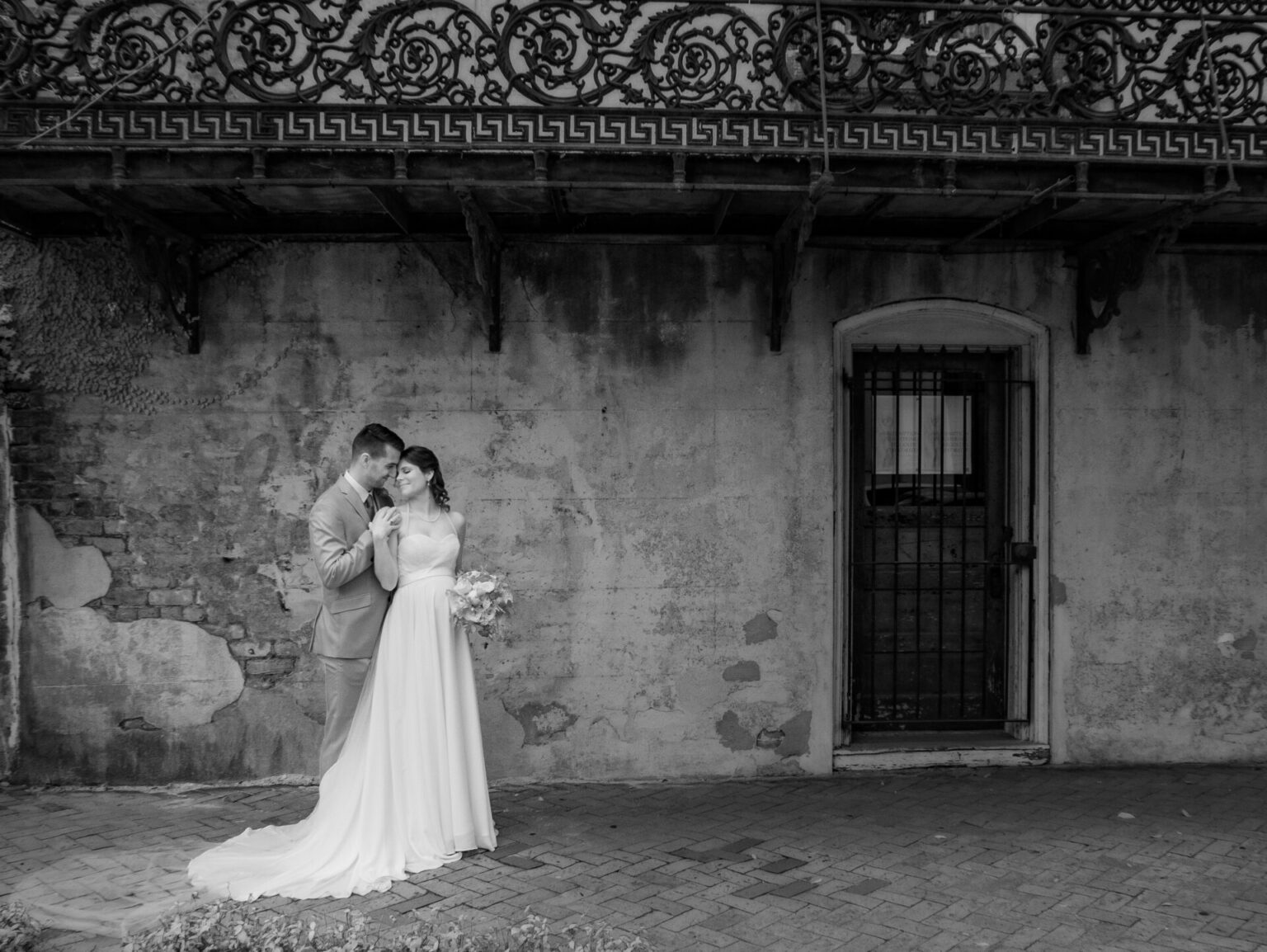 Elopement Wedding A bride and groom stand closely under a wrought iron balcony in a black and white photo. The bride wears a long, flowing gown and holds a bouquet, while the groom in a suit gazes at her. They're positioned on a cobblestone path beside a textured, weathered wall with a barred door. Elopements Inc