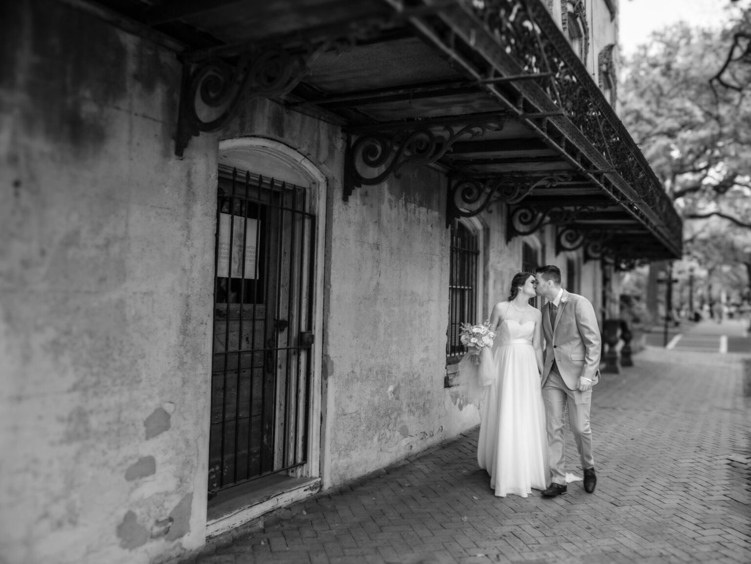 Elopement Wedding A black-and-white photo of a couple on their wedding day. The bride, in a flowing gown, holds a bouquet. The groom wears a suit. They share a kiss while standing on a brick sidewalk under an ornate iron balcony. The building's aged facade adds a vintage charm to the scene. Elopements Inc