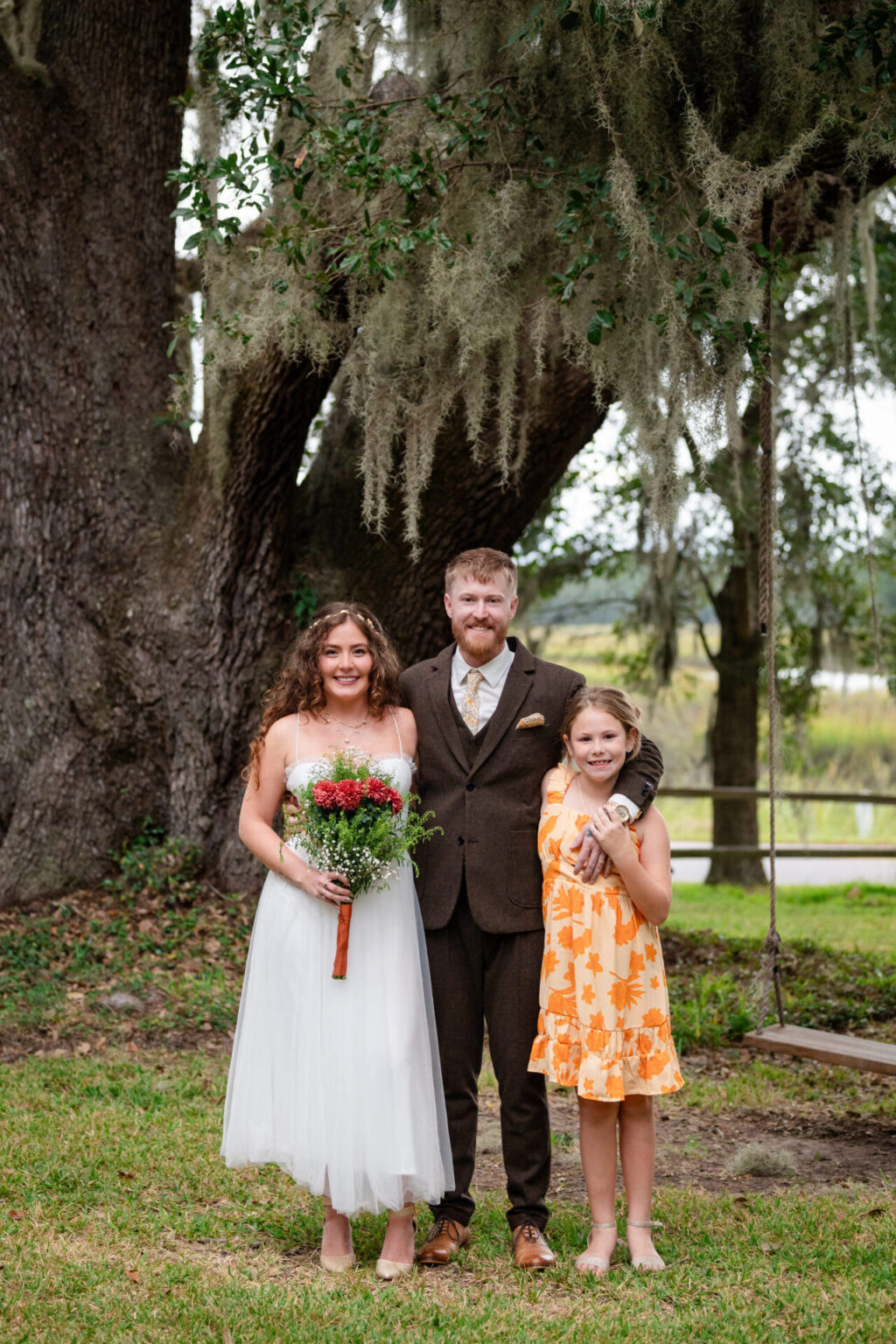 Elopement Wedding A bride in a white dress holds a bouquet of red and white flowers, standing beside a groom in a brown suit. A young girl in an orange floral dress stands next to them. They are outdoors under a large tree with hanging moss. A swing and grassy area are visible in the background. Elopements Inc