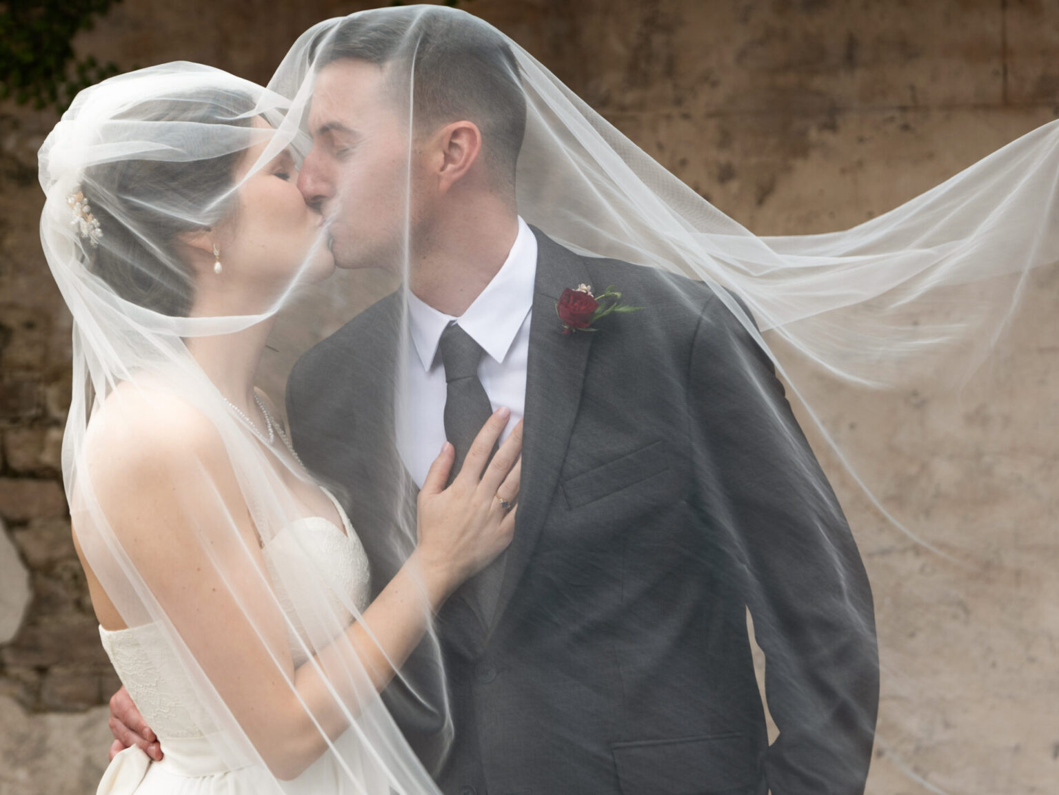 Elopement Wedding A bride and groom kiss under a sheer veil. The bride, in a strapless white gown, has her hair styled with a hairpiece. The groom wears a dark suit with a white shirt and a rose boutonniere. The veil gently flows in the wind against a textured, rustic background. Elopements Inc