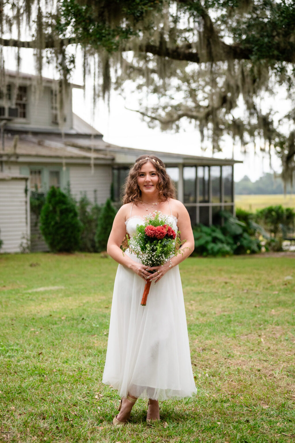 Elopement Wedding A woman in a white dress stands on a grassy lawn holding a bouquet of red and white flowers. Her curly hair cascades down her shoulders. In the background, there's a house with hanging moss from nearby trees. The setting appears serene and outdoorsy. Elopements Inc