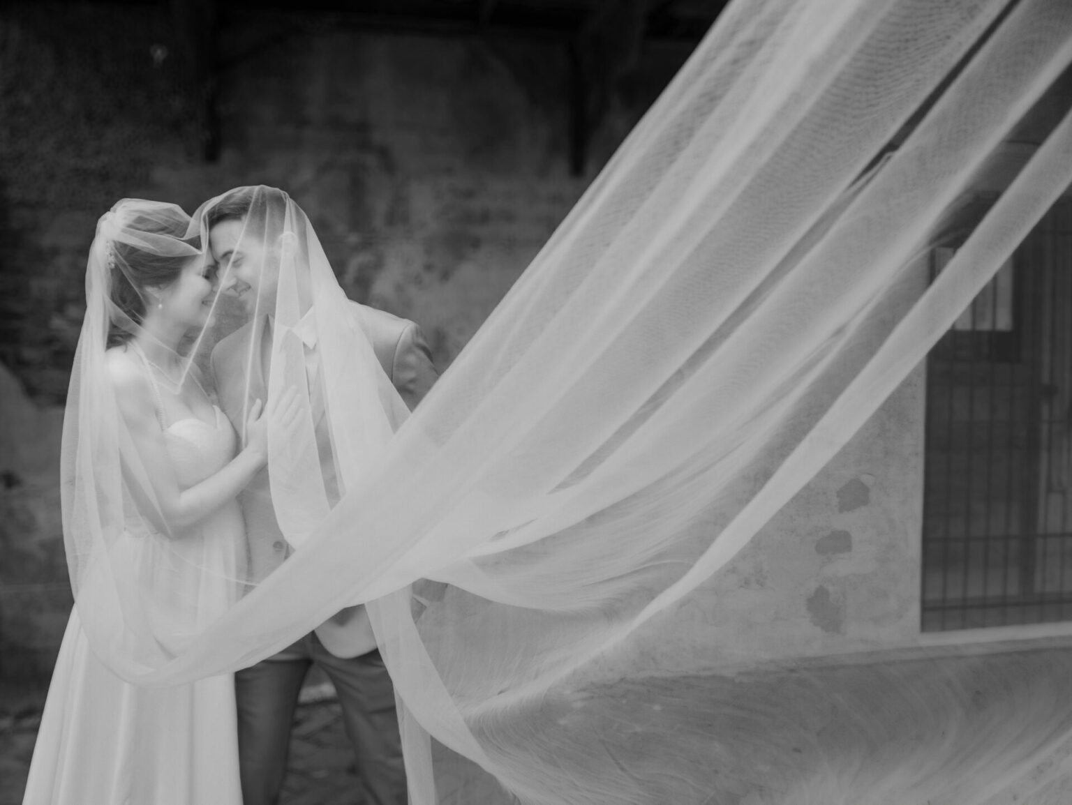 Elopement Wedding A black and white photo of a newlywed couple embracing under a long, flowing veil. The bride wears a strapless gown, and the groom is in a suit. They stand closely, smiling and touching foreheads, with the veil billowing dramatically in the foreground, creating a romantic scene. Elopements Inc