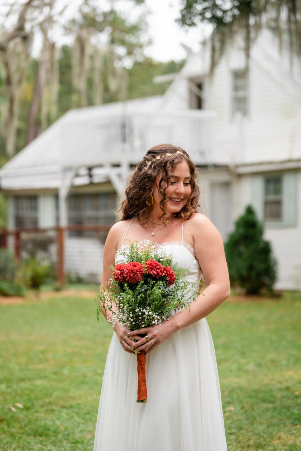 Elopement Wedding A bride wearing a white dress, holding a bouquet of red and white flowers, smiles while standing on a grassy lawn. Her curly hair is adorned with small flowers. A white house with large windows and a porch is blurred in the background, surrounded by greenery. Elopements Inc