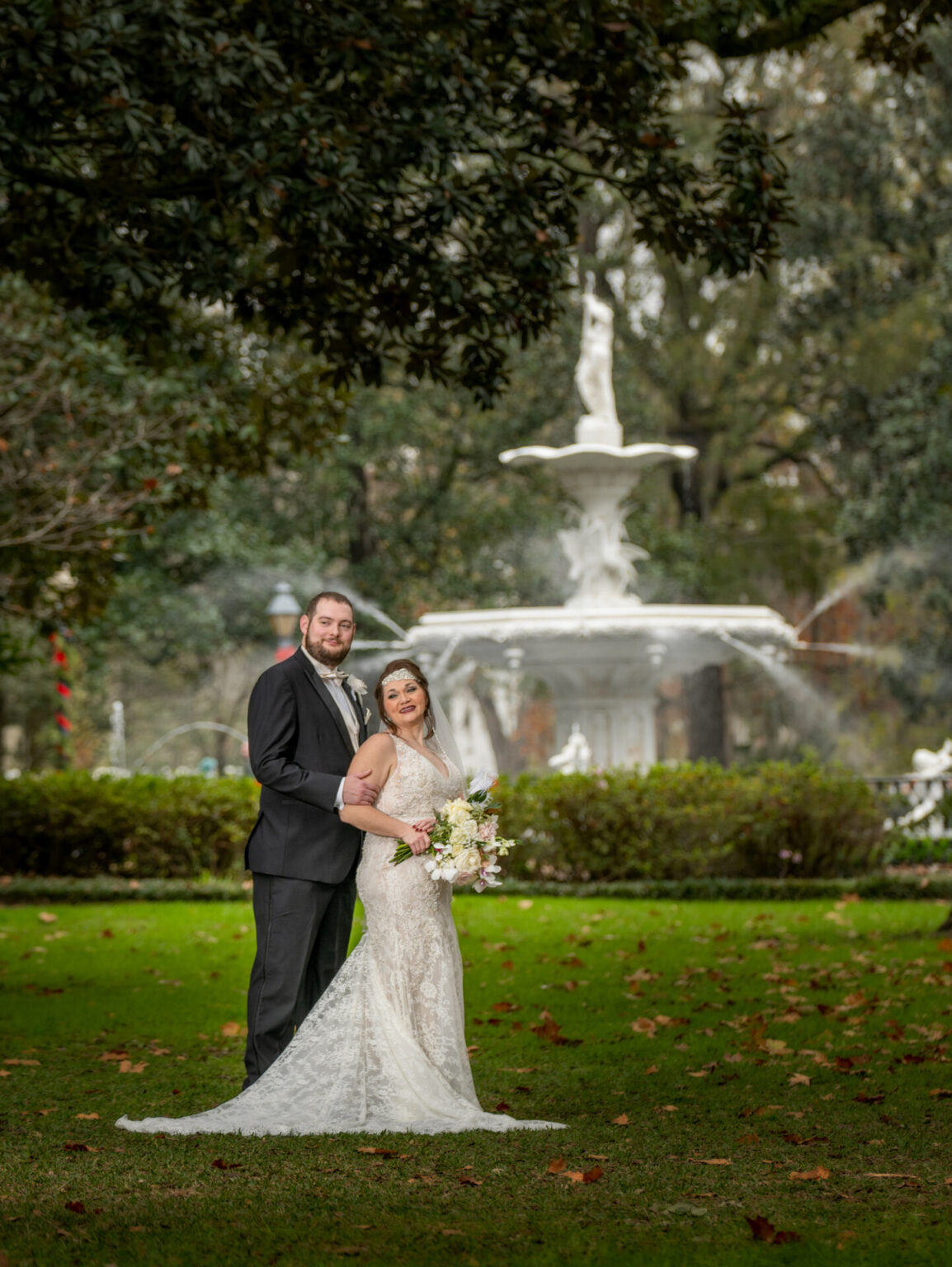 Elopement Wedding A bride and groom stand together on a green lawn in front of a large white fountain. The bride is in a lace wedding dress, holding a bouquet, and the groom is in a black suit. The park setting includes lush trees and scattered leaves, creating a serene atmosphere. Elopements Inc