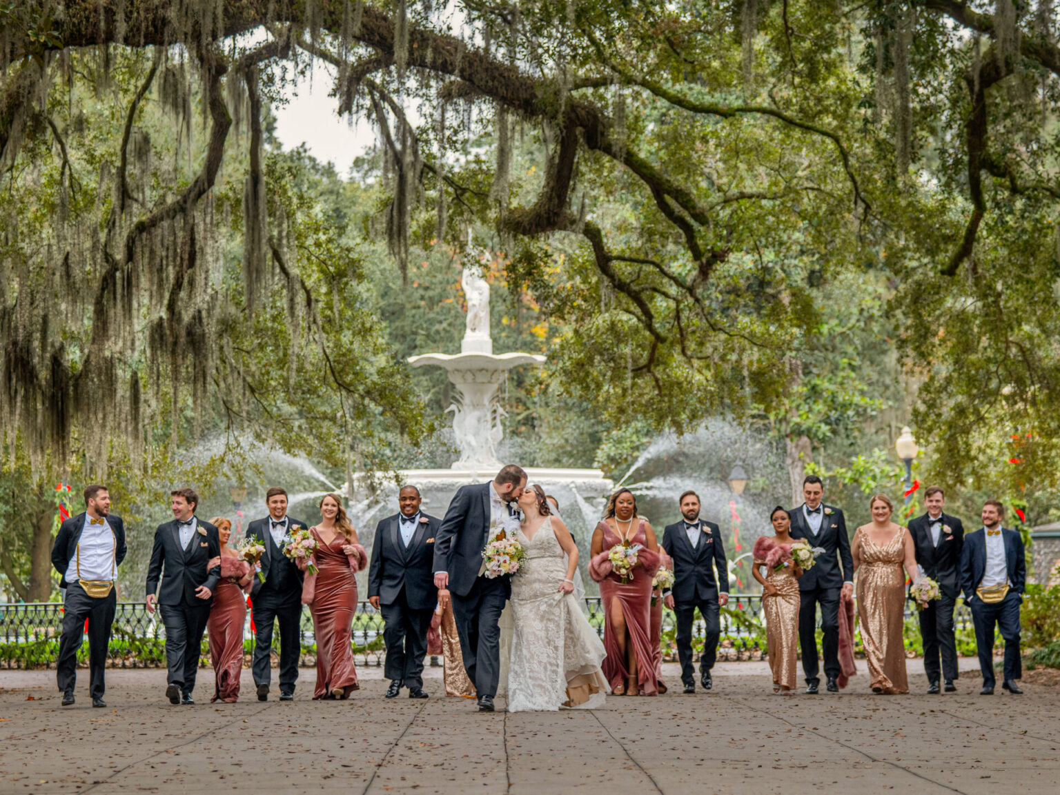 Elopement Wedding A wedding party walks toward the camera under sprawling oak trees with Spanish moss. The bride and groom are in the center, with bridesmaids in burgundy dresses and groomsmen in black suits on either side. A white fountain is in the background, set against a lush, green park setting. Elopements Inc