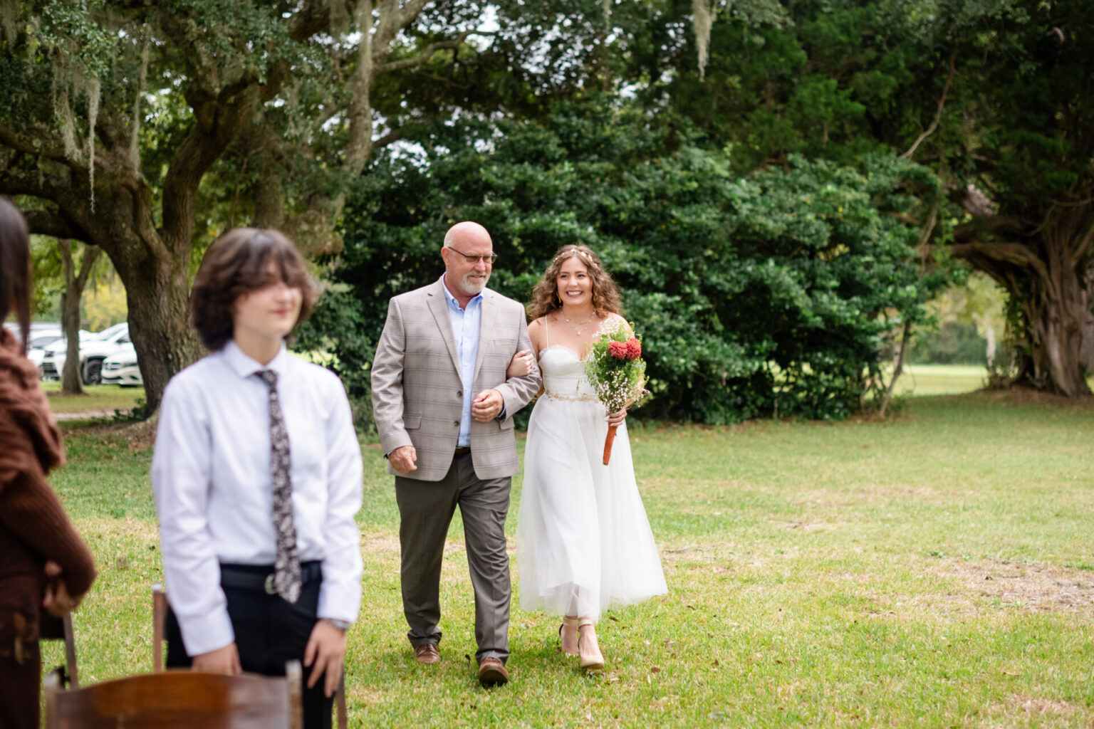 Elopement Wedding A bride in a white off-shoulder dress walks arm in arm with an older man in a gray suit, smiling. She holds a bouquet of red and white flowers. They walk through a grassy area with large trees. A young man in a white shirt and tie stands nearby, with chairs and guests in the foreground. Elopements Inc