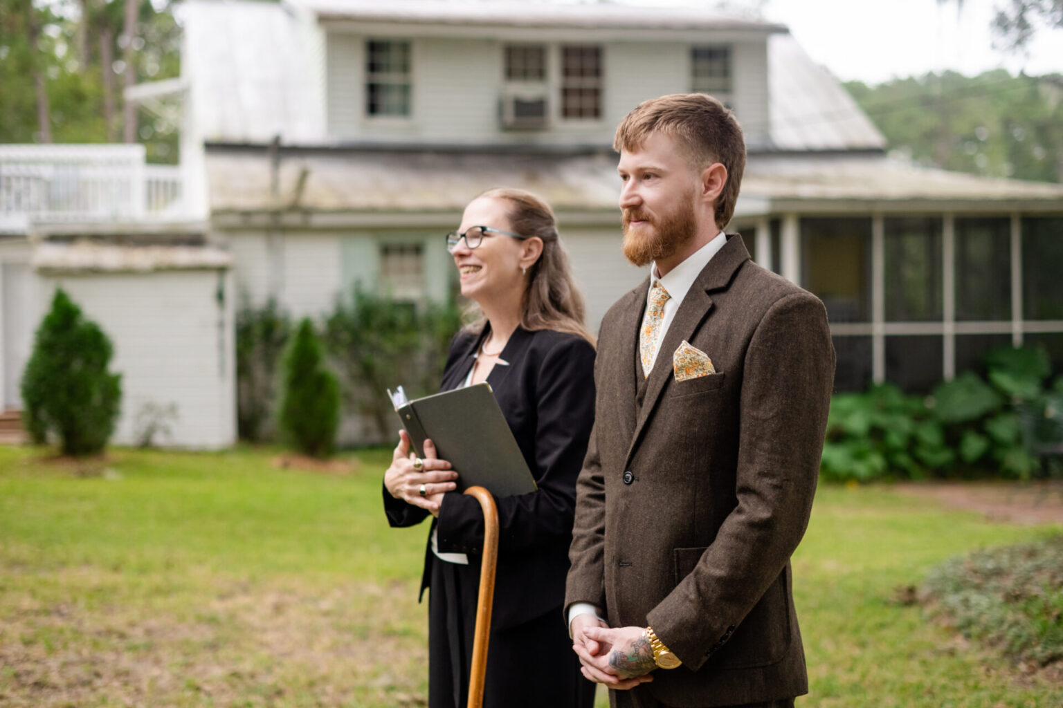 Elopement Wedding A man in a brown suit and patterned tie stands beside a smiling woman holding a notebook. They are outdoors on a grassy area with bushes and a white two-story house in the background. The man has a beard and the woman wears glasses and is dressed in black. Elopements Inc