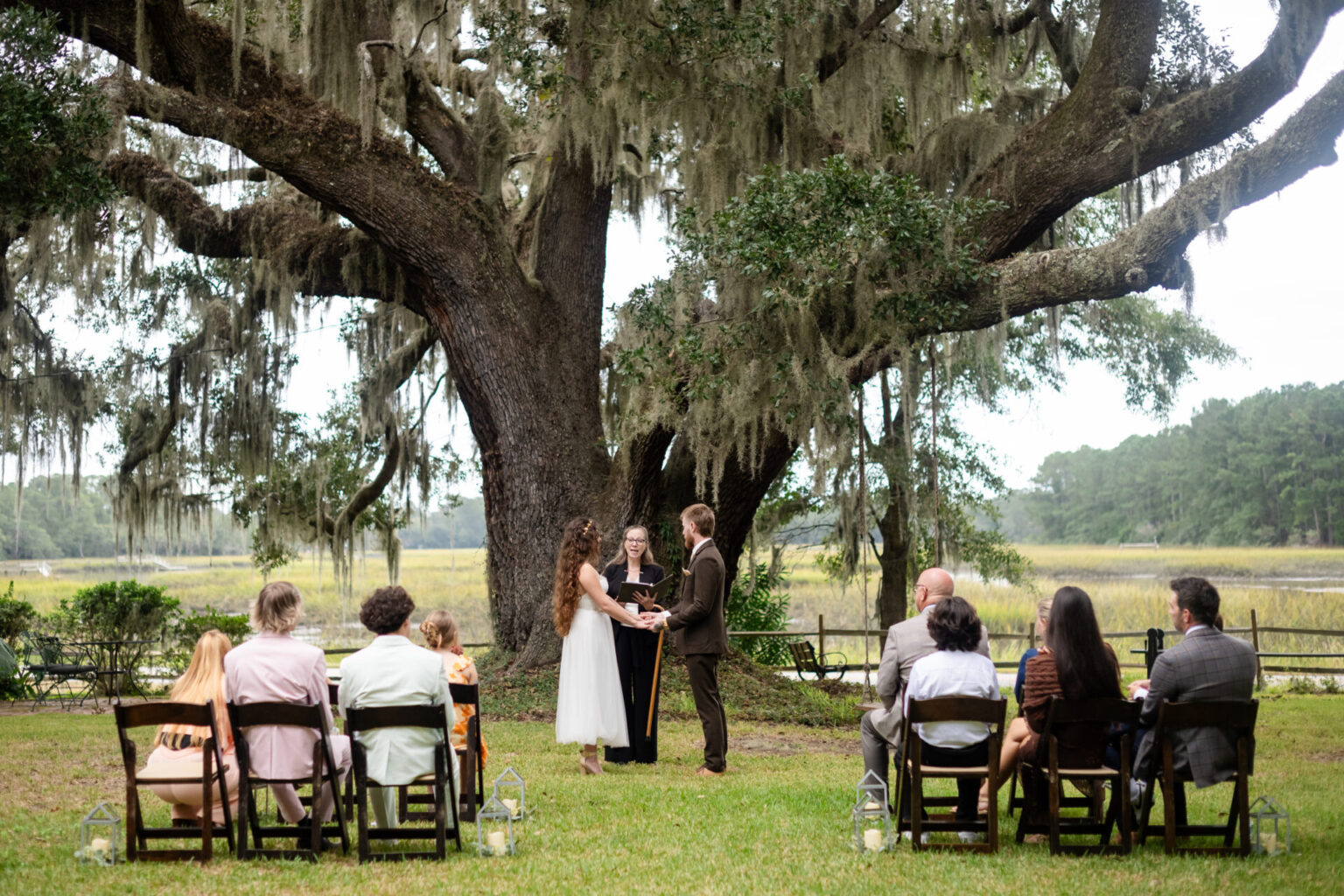 Elopement Wedding A wedding ceremony is taking place outdoors under a large, moss-covered tree. The couple stands facing each other, holding hands, with an officiant between them. Guests are seated on wooden chairs in rows, wearing semi-formal attire. A scenic landscape with grass and trees is in the background. Elopements Inc