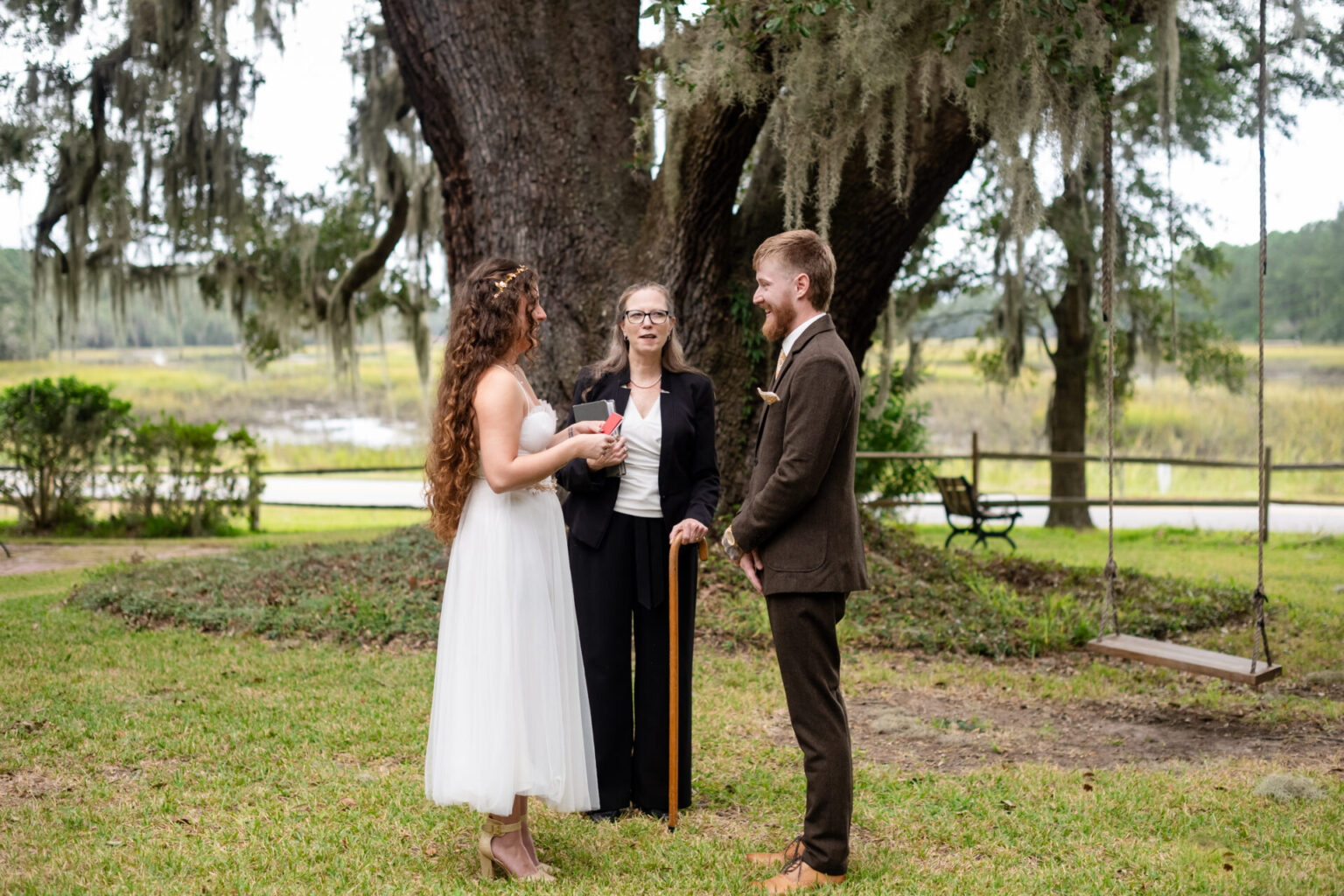 Elopement Wedding A bride in a white dress and a groom in a brown suit stand facing each other during an outdoor wedding ceremony. An officiant in black holds a book and stands between them. They are beneath a large tree with sprawling branches, and moss hangs down. A grassy area and wooden bench are nearby. Elopements Inc