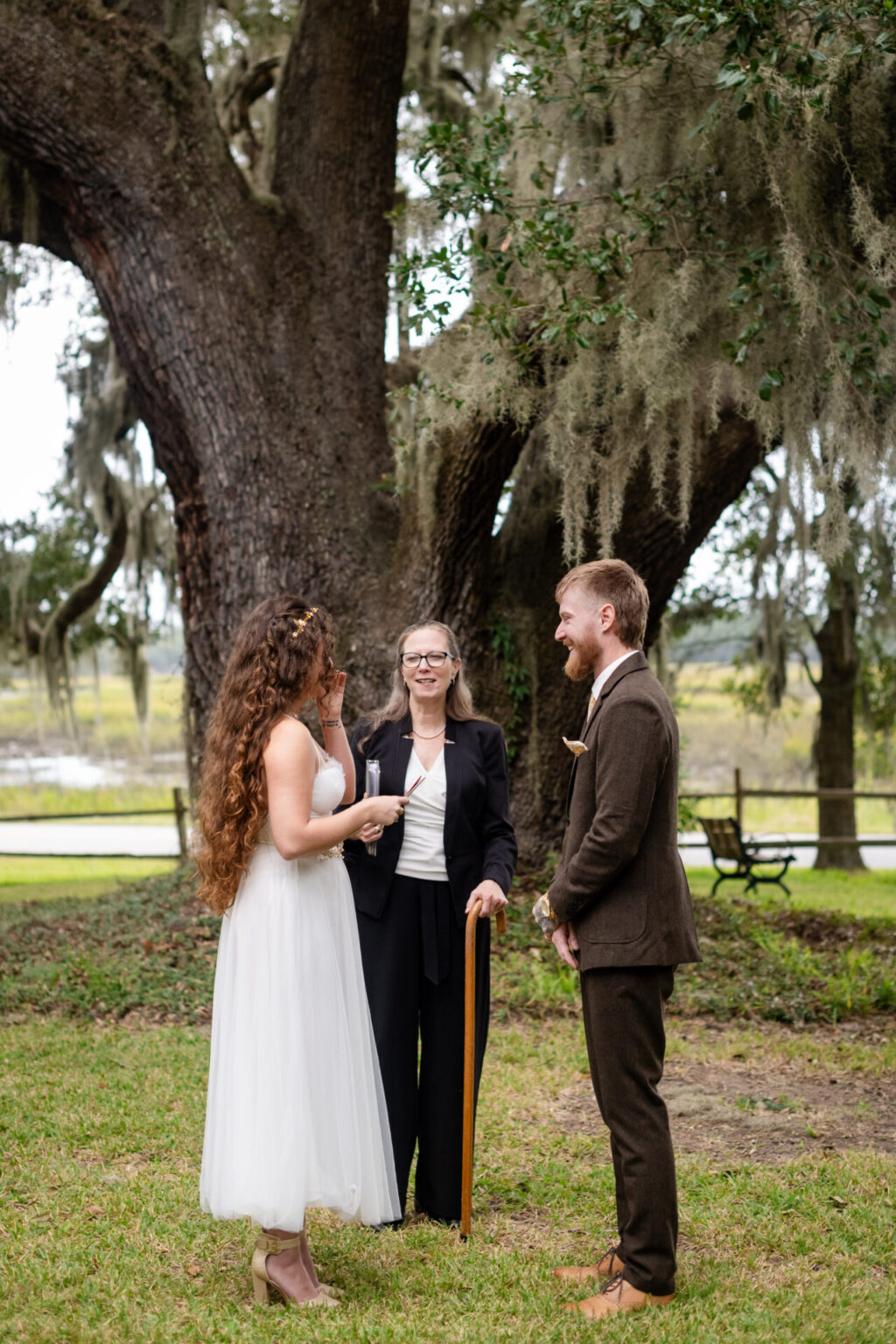 Elopement Wedding A bride in a white dress and groom in a brown suit smile at each other during their outdoor wedding ceremony. An officiant with long hair stands between them, holding a microphone. They are beneath a large, moss-covered tree, with a wooden fence and grassy field in the background. Elopements Inc