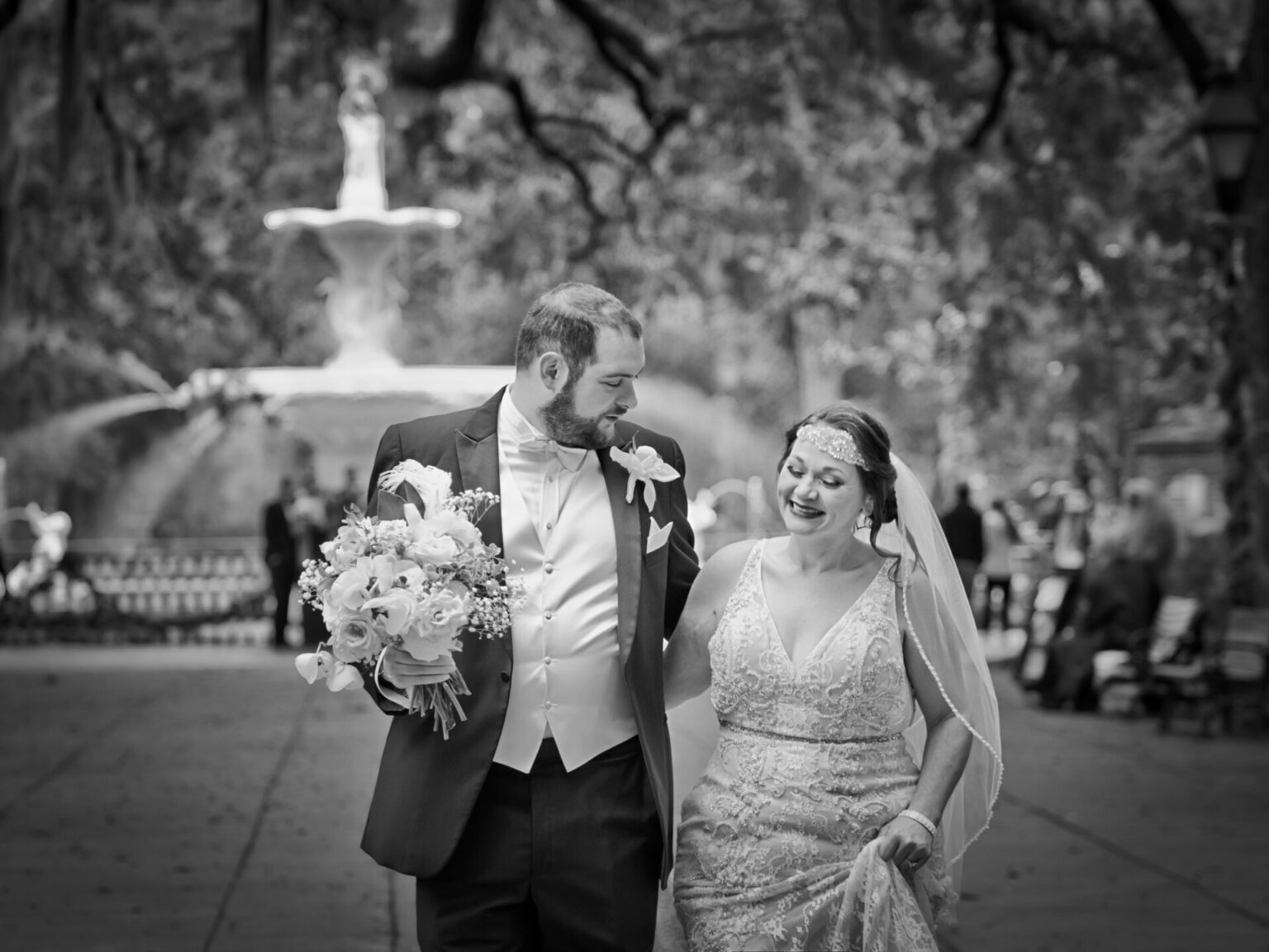 Elopement Wedding A joyful couple walks hand in hand down a tree-lined path in a park. The groom, in a dark suit with a boutonniere, carries a floral bouquet. The bride, in a lace gown and veil, smiles warmly. A fountain is in the blurred background. The image is in black and white, enhancing its timeless feel. Elopements Inc