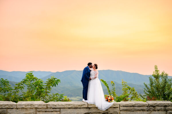 Elopement Wedding A couple in wedding attire stands on a stone wall against a backdrop of mountains. The bride, in a white gown, and the groom, in a blue suit, kiss under a vibrant orange sky. Lush greenery surrounds them, and there is a colorful bouquet at their feet. Elopements Inc