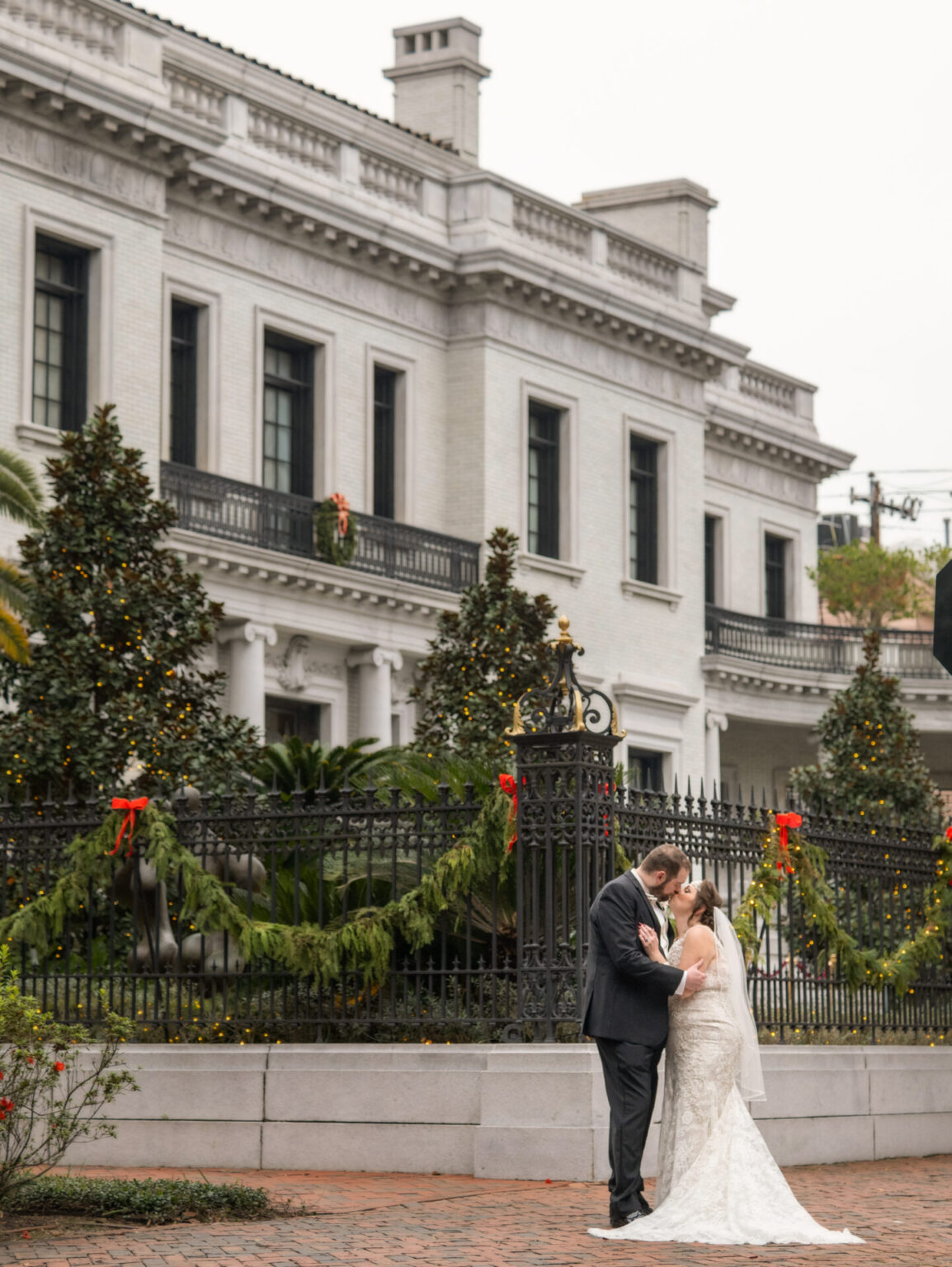 Elopement Wedding A bride and groom share an embrace in front of a large, ornate white building with tall windows and a black iron fence adorned with festive garlands and red bows. The bride wears a lace gown and veil, and the groom is in a dark suit. Trees and shrubs are visible in the background. Elopements Inc