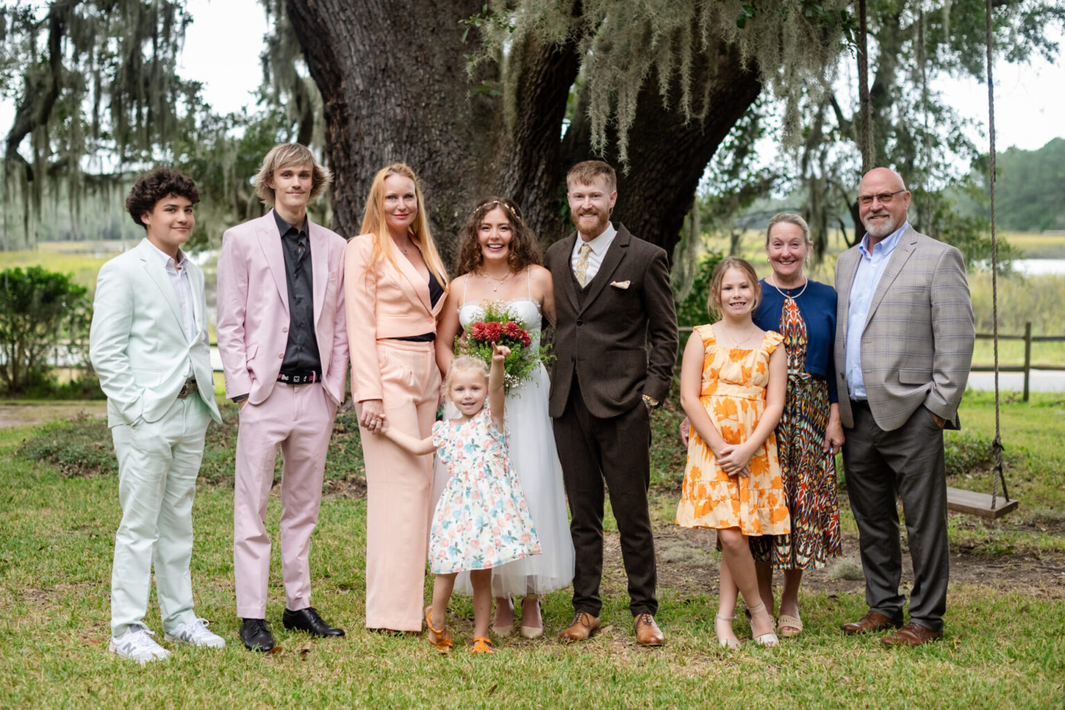 Elopement Wedding A group photo of eight people posing outdoors under a large tree. The couple in the center is dressed in wedding attire—she in a white gown holding a bouquet and he in a brown suit. Others wear various colorful outfits, standing on grass, with a fence and trees in the background. Elopements Inc