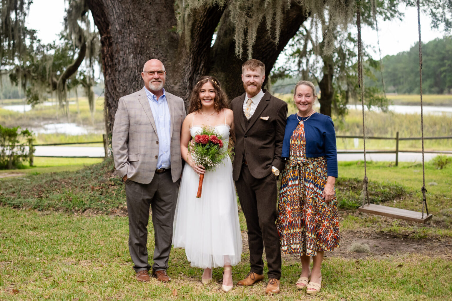 Elopement Wedding A bride and groom stand smiling under a large tree with moss, flanked by two adults. The bride holds a bouquet and wears a white dress, while the groom dons a brown suit. The man to the left wears a plaid blazer, and the woman on the right wears a colorful dress. A swing hangs nearby. Elopements Inc