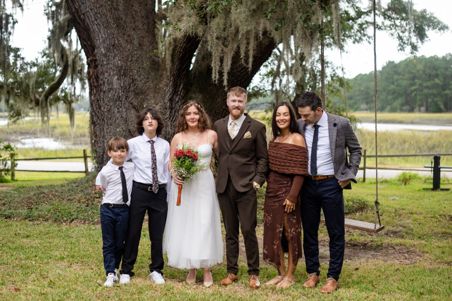 Elopement Wedding A wedding party poses outdoors. A bride in a white dress holds flowers, and a groom in a brown suit stands beside her. Two men in dark suits, a woman in a brown dress, and a young boy in a white shirt and tie are beside them. They're in front of a large tree with moss and a swing. Elopements Inc