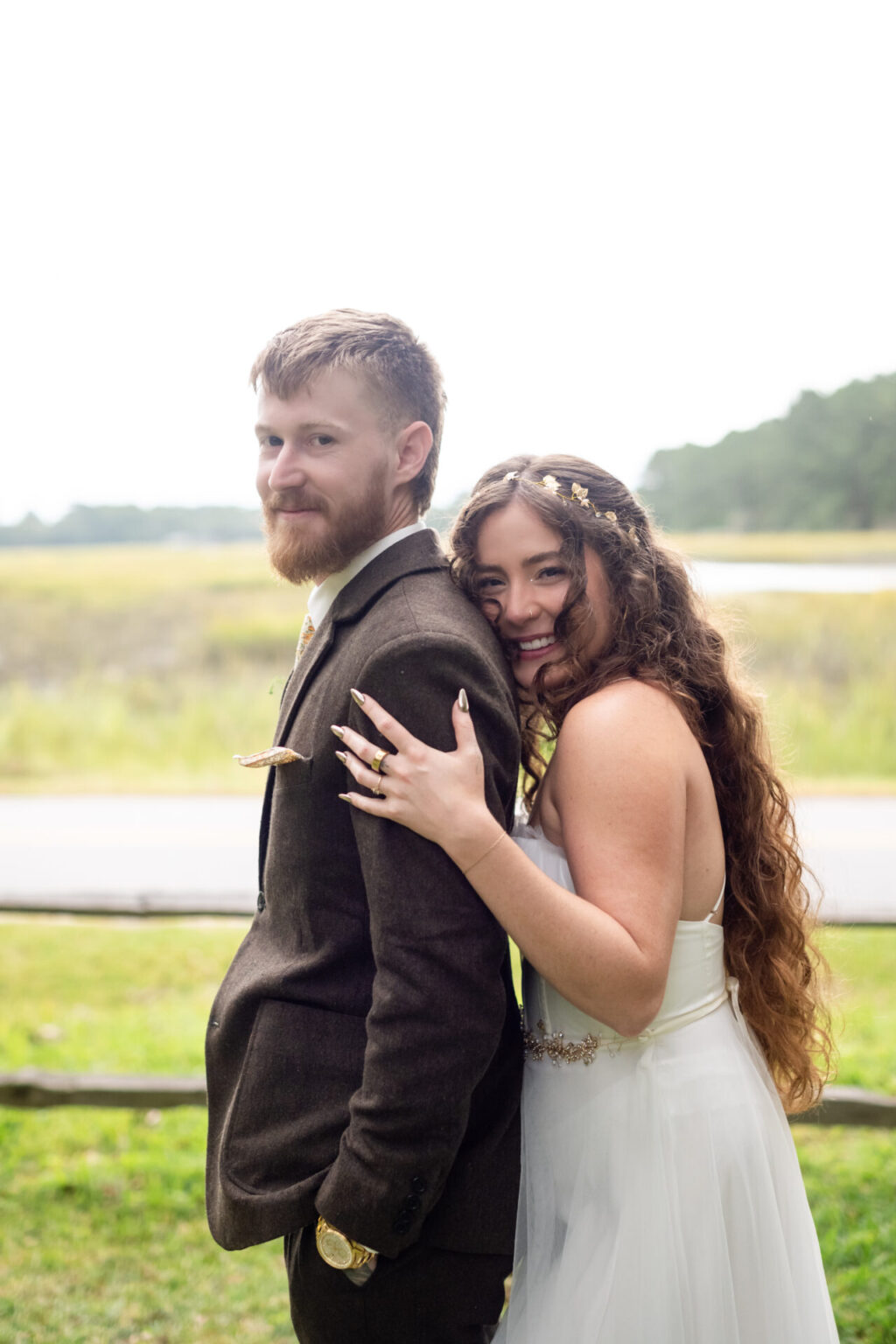 Elopement Wedding A smiling couple poses outdoors. The man in a brown suit has a beard and looks at the camera. The woman, with long curly hair, rests her hand with decorated nails on his shoulder, wearing a white dress. A wooden fence and greenery are visible in the background under a bright sky. Elopements Inc