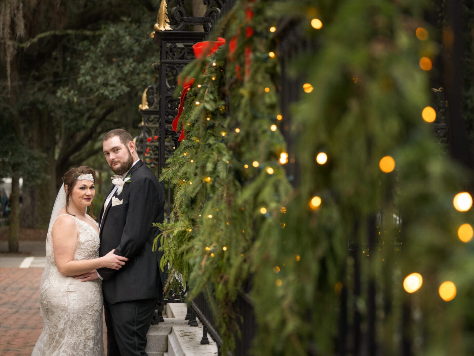 Elopement Wedding A bride and groom stand close together by an ornate wrought iron fence adorned with evergreen garlands and string lights. The bride wears a lace dress and headpiece with a veil, while the groom is in a black suit. The festive decorations suggest a holiday theme. Elopements Inc