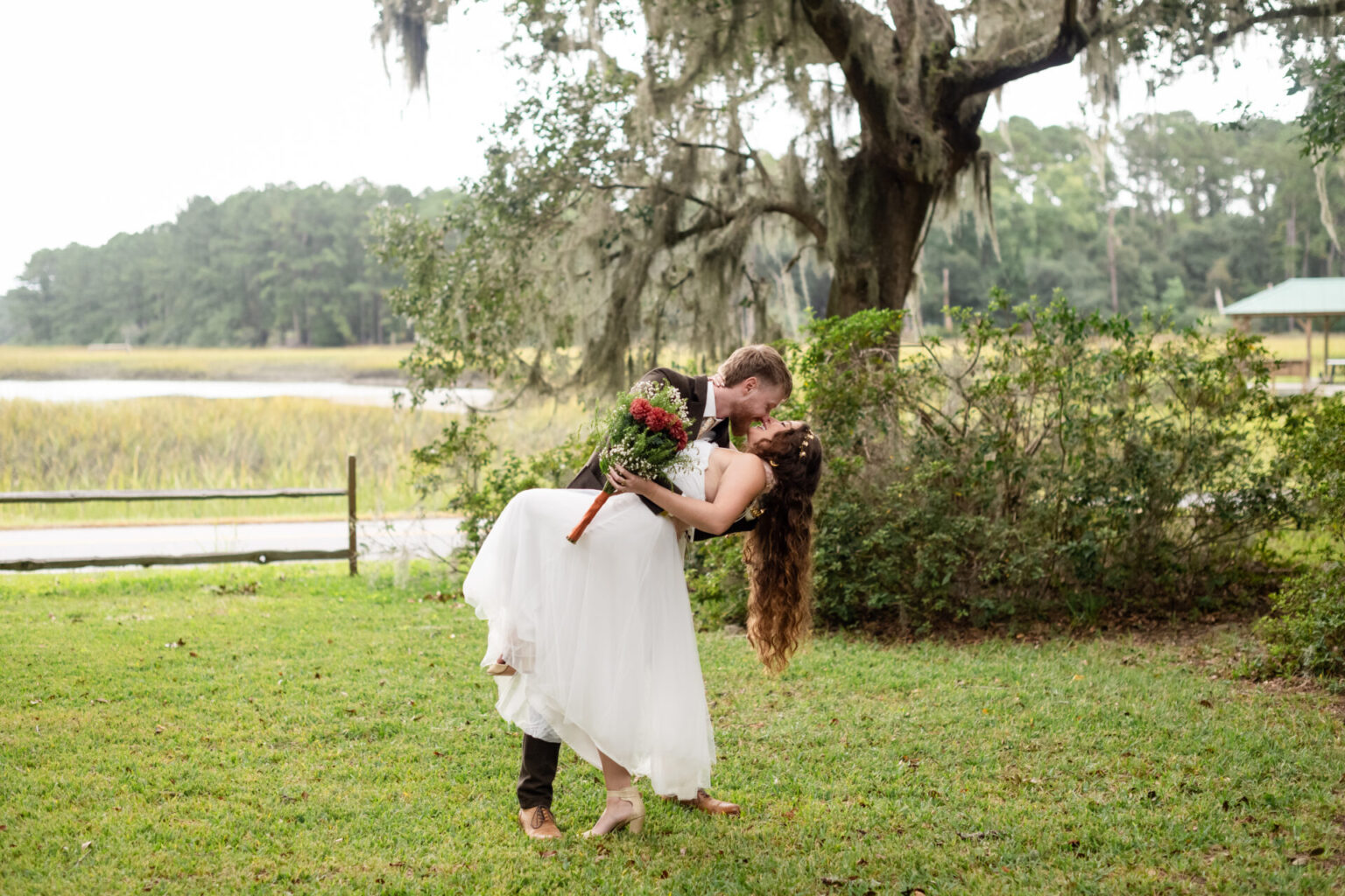 Elopement Wedding A couple in formal attire shares a romantic kiss outdoors. The man, in dark pants and a white shirt, leans the woman back. She wears a white dress and holds a bouquet of flowers. They stand on a grassy area with trees and a wooden fence in the background, under an overcast sky. Elopements Inc