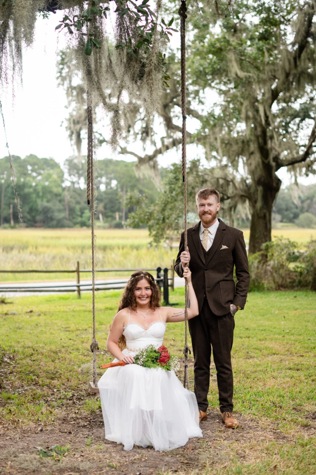 Elopement Wedding A bride in a white dress sits on a rope swing holding a bouquet with red and white flowers. A groom in a dark suit stands beside her, holding the swing's rope. They're outdoors with a grassy field, large oak tree, and wooden fence in the background. Moss hangs from the tree branches. Elopements Inc