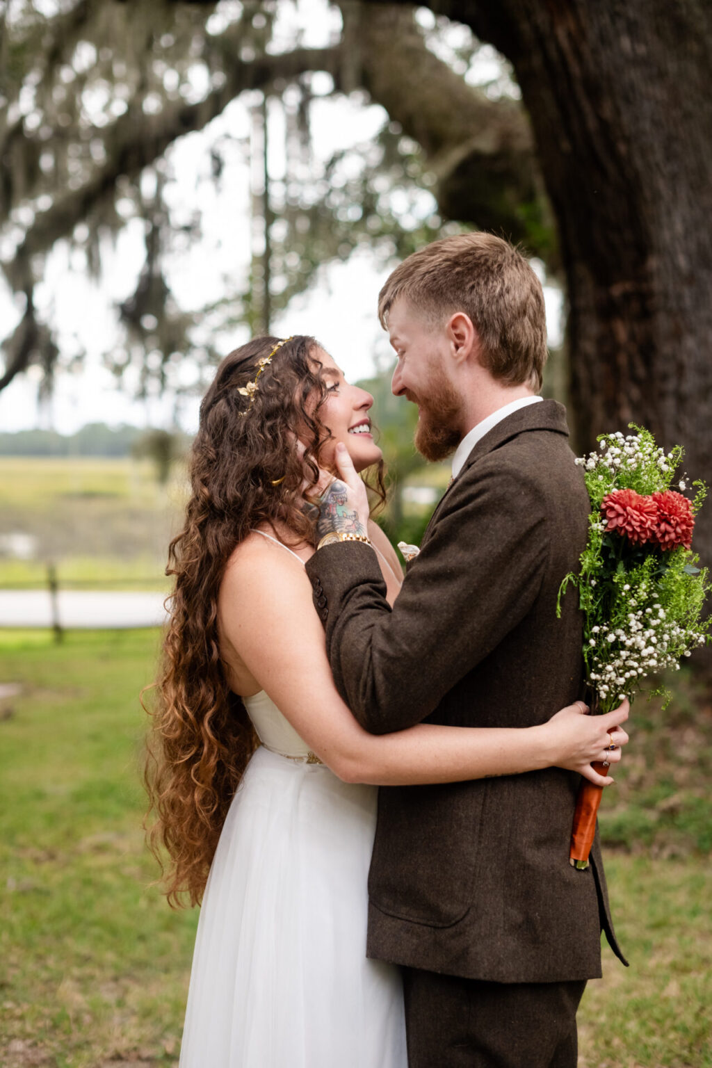 Elopement Wedding A smiling bride and groom embrace outdoors. The bride, with long curly hair, wears a white dress. The groom, with a beard, wears a brown suit. She holds a bouquet of red and white flowers. A large tree and a wooden fence are visible in the background, suggesting a rustic setting. Elopements Inc