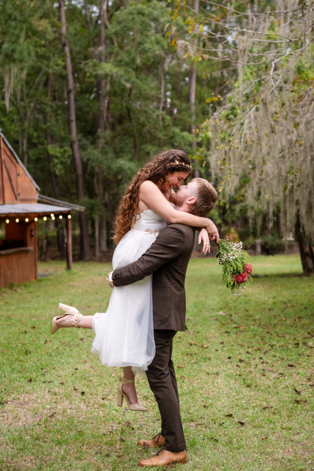 Elopement Wedding A groom lifts a bride in a white dress as they kiss outdoors. The bride holds a bouquet of red and white flowers. They are in a grassy area with tall trees and a wooden building in the background. The groom is in a dark suit, and both are smiling with joy. Elopements Inc