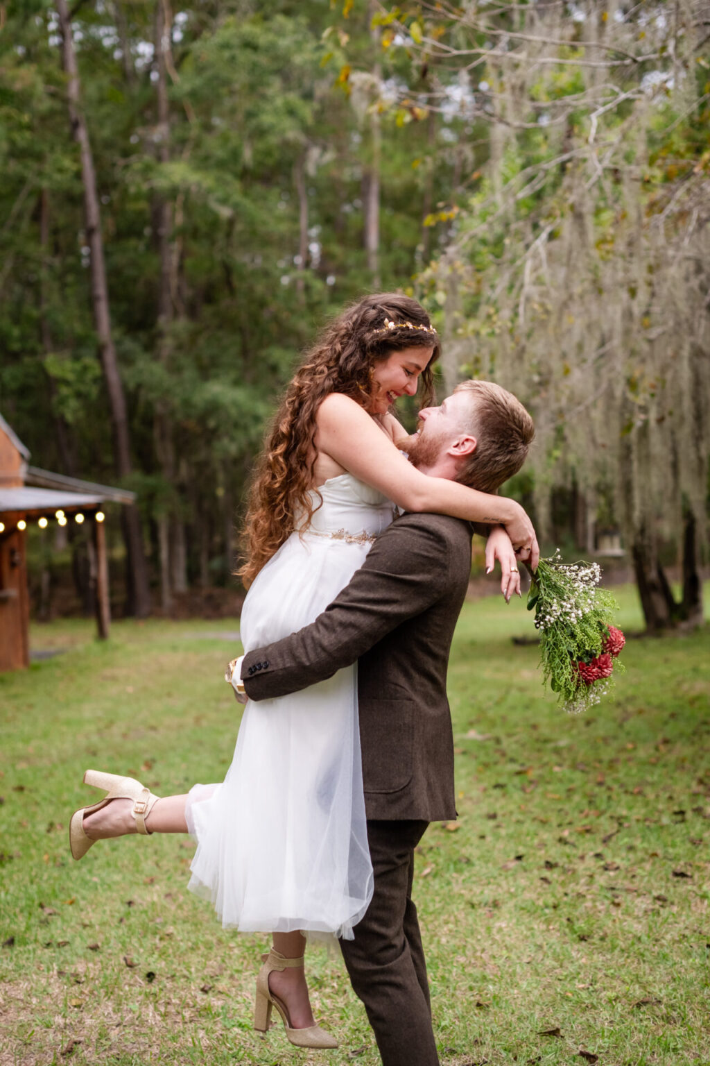 Elopement Wedding A bride in a white dress with flowing, wavy hair and floral crown is joyfully lifted by a groom in a brown suit. She's holding a bouquet with red and white flowers. They are in a wooded outdoor setting with a small wooden building in the background, surrounded by green grass and trees. Elopements Inc