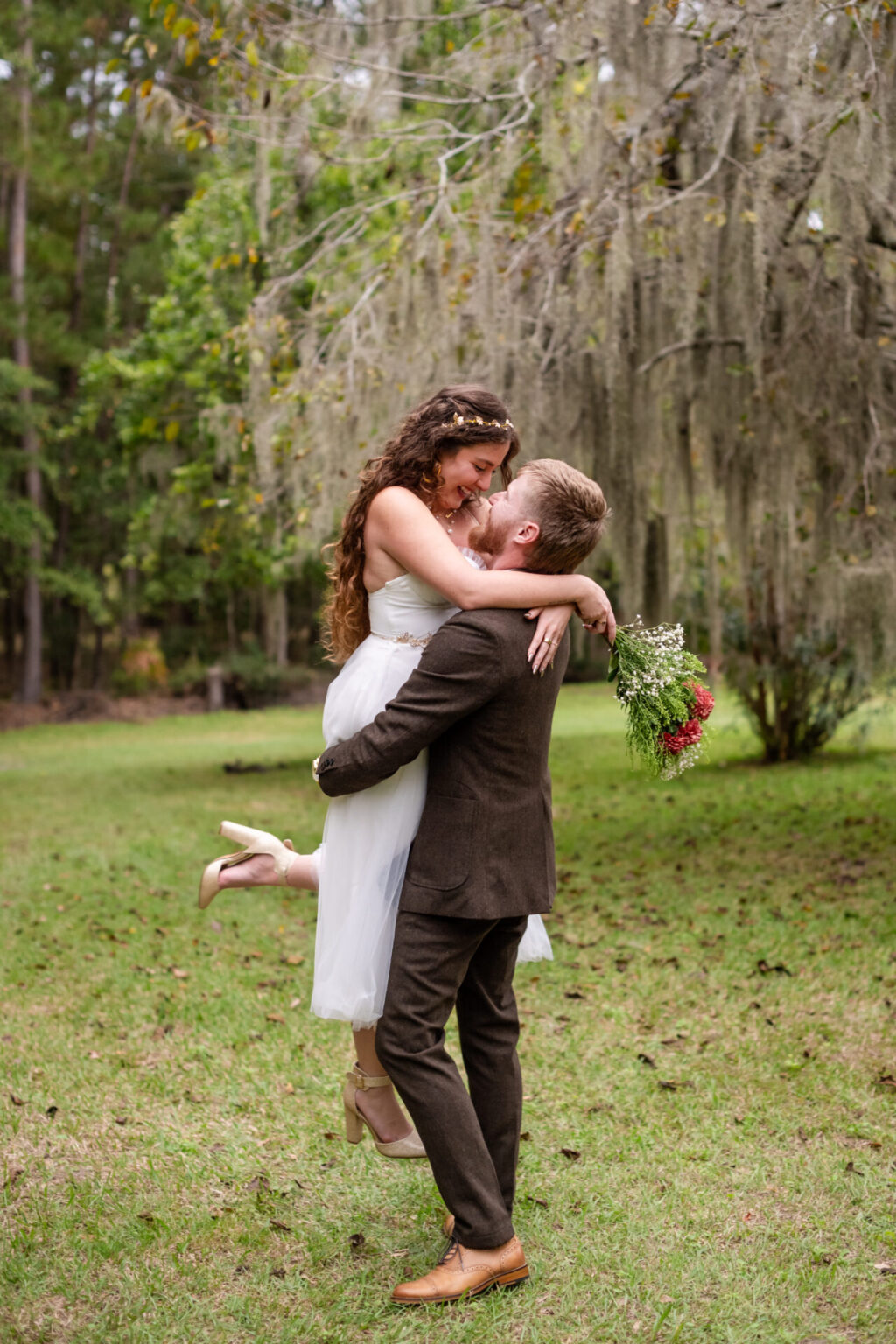 Elopement Wedding A couple is celebrating outdoors, where a man in a brown suit lifts a woman in a white dress, who holds a bouquet of flowers. They are in a grassy area surrounded by trees draped with Spanish moss. Both are smiling and appear joyful. Elopements Inc