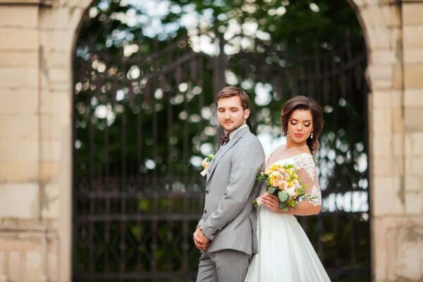 Elopement Wedding A bride and groom stand back-to-back in front of an ornate iron gate. The groom wears a gray suit and the bride wears a white gown with lace sleeves, holding a bouquet of yellow and orange flowers. Both have closed eyes, set against a blurred background of greenery. Elopements Inc